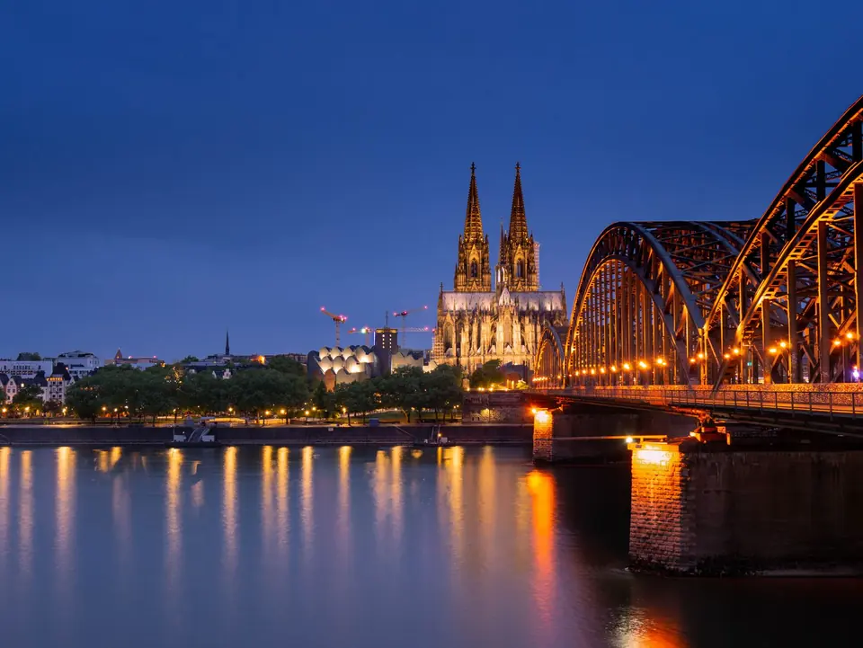 Der ikonische Kölner Dom und die Hohenzollernbrücke wunderschön beleuchtet gegen den Nachthimmel, der sich im Rhein spiegelt.